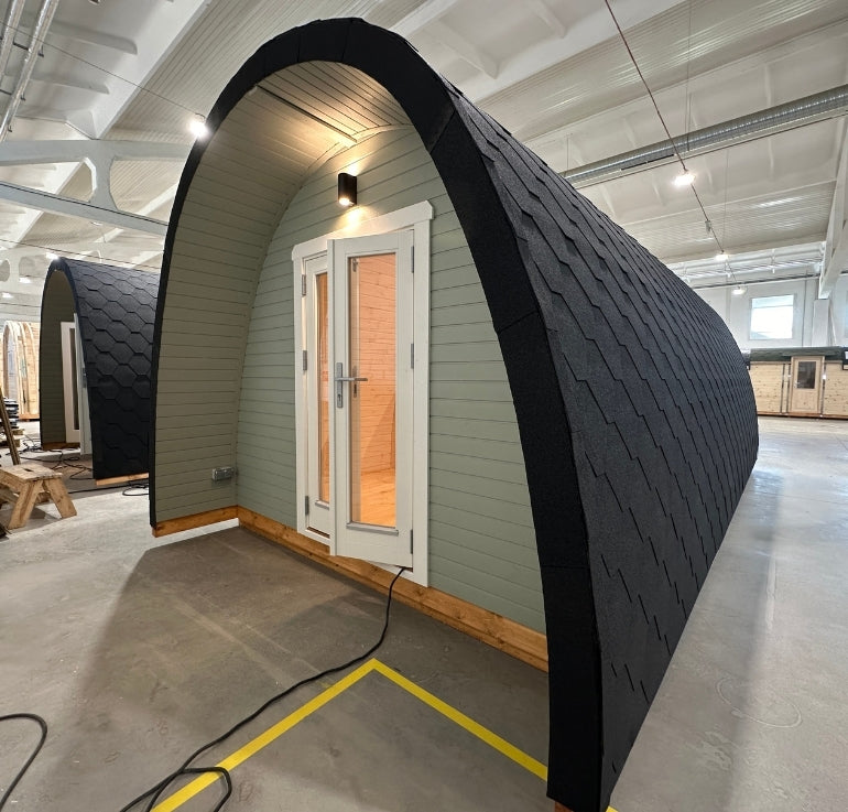 Interior view of the GlampLaunch factory showing a luxury pod mid-production, with full cladding, glass doors, and shingle roof under industrial lighting.