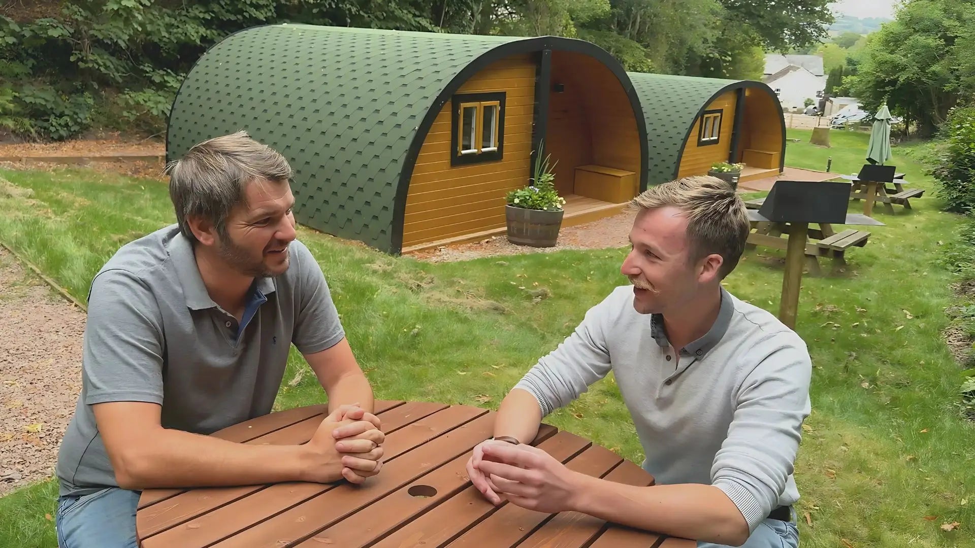 Two men sitting at a wooden table in front of green and yellow pod-style cabins.