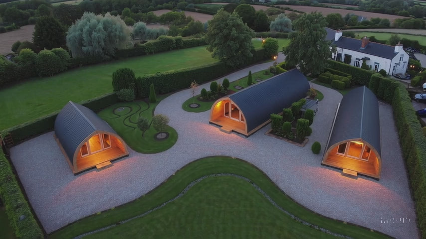 Aerial view of three curved timber glamping pods spaced apart on a landscaped private site beside a countryside home, with gravel paths, decks, and outdoor seating.