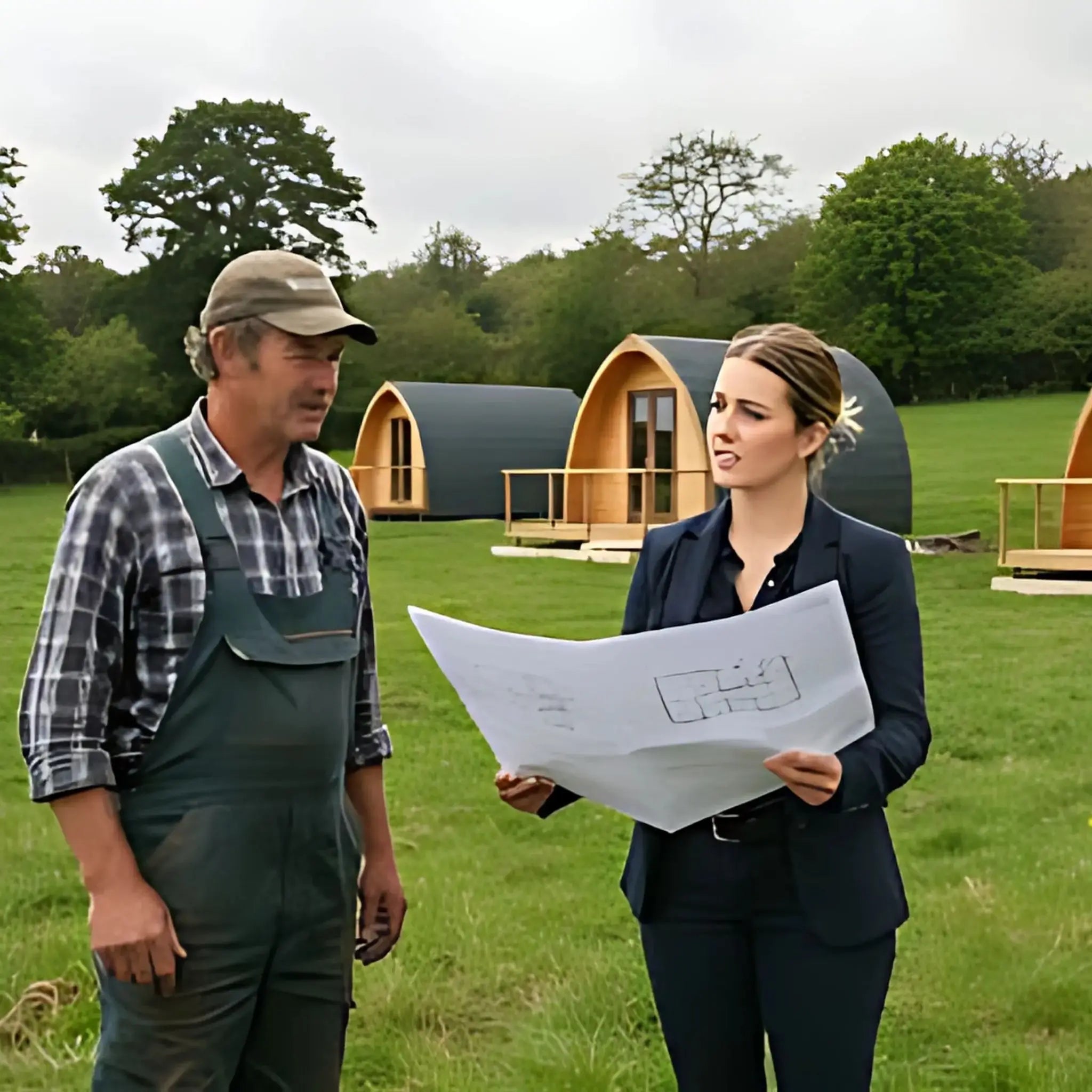 A farmer and a business consultant reviewing plans for a glamping site, with newly installed wooden pods in the background of a grassy field.