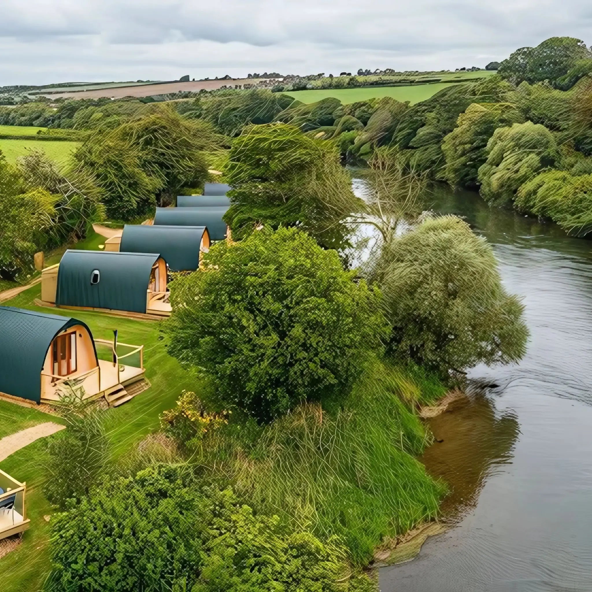 A row of luxury glamping pods with green roofs overlooking a tranquil river and lush countryside.