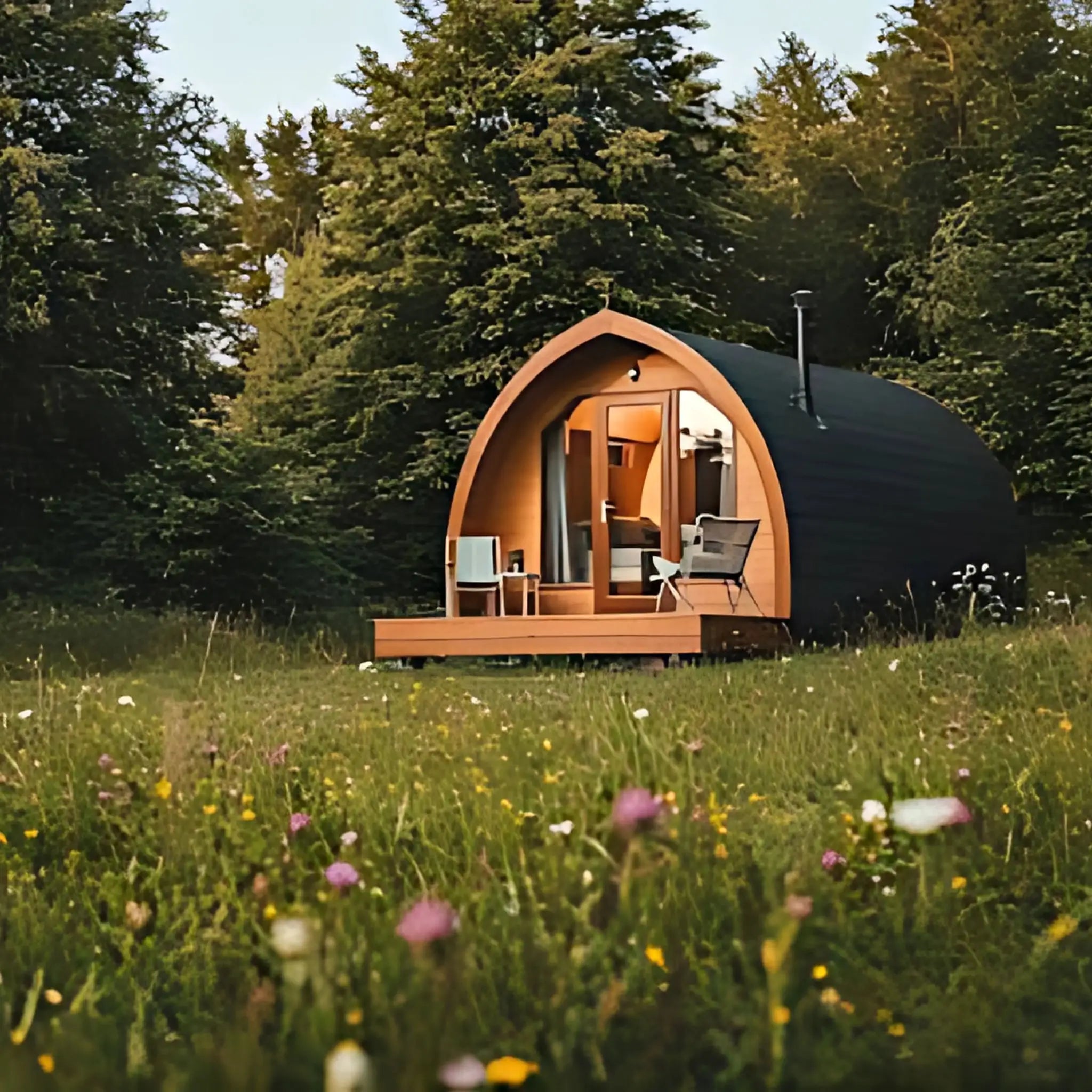A cosy wooden glamping pod with a deck and seating area, nestled in a quiet forest clearing surrounded by wildflowers.