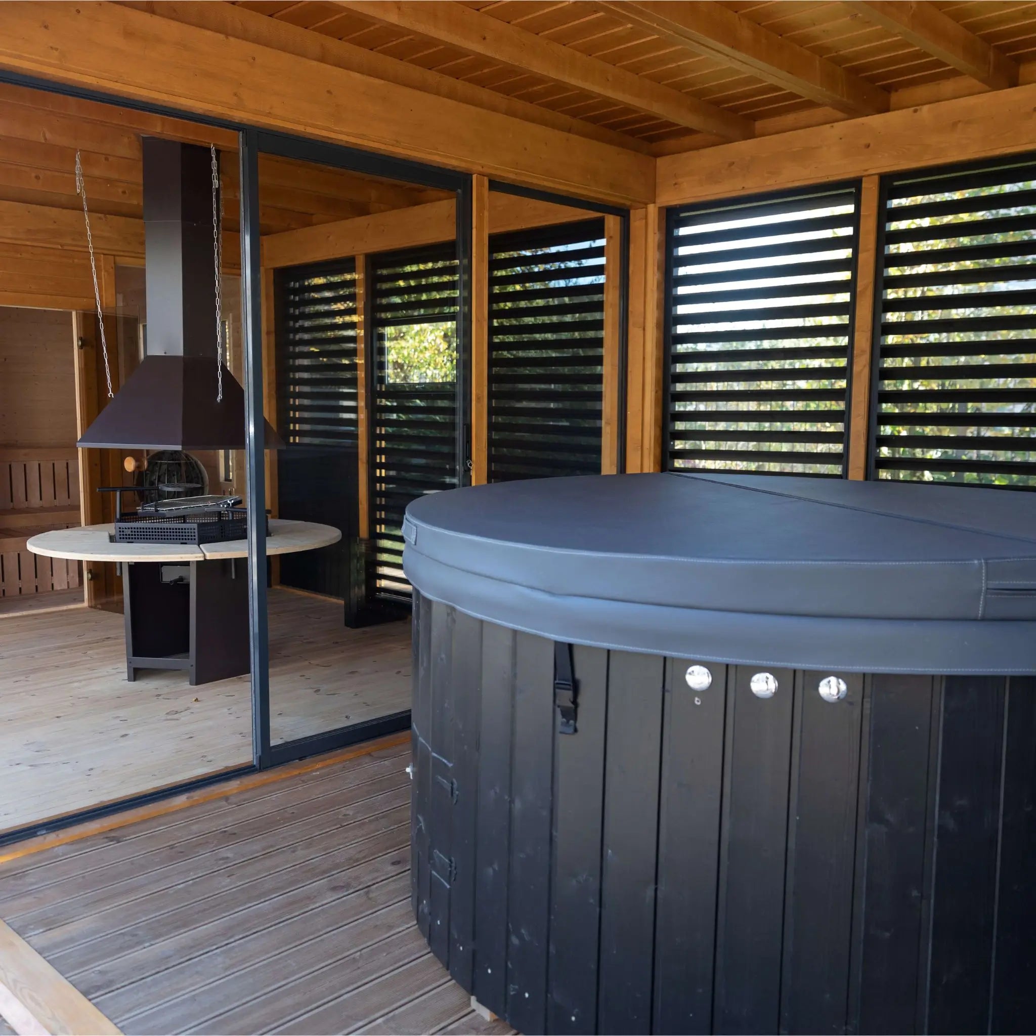 Interior view of a luxury spa garden cube featuring a covered hot tub and modern grill area, separated by louvered glass panels for a seamless wellness experience.