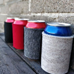 Wool can coolers holding assorted drink cans on a dark wooden bench beside a stone wall.