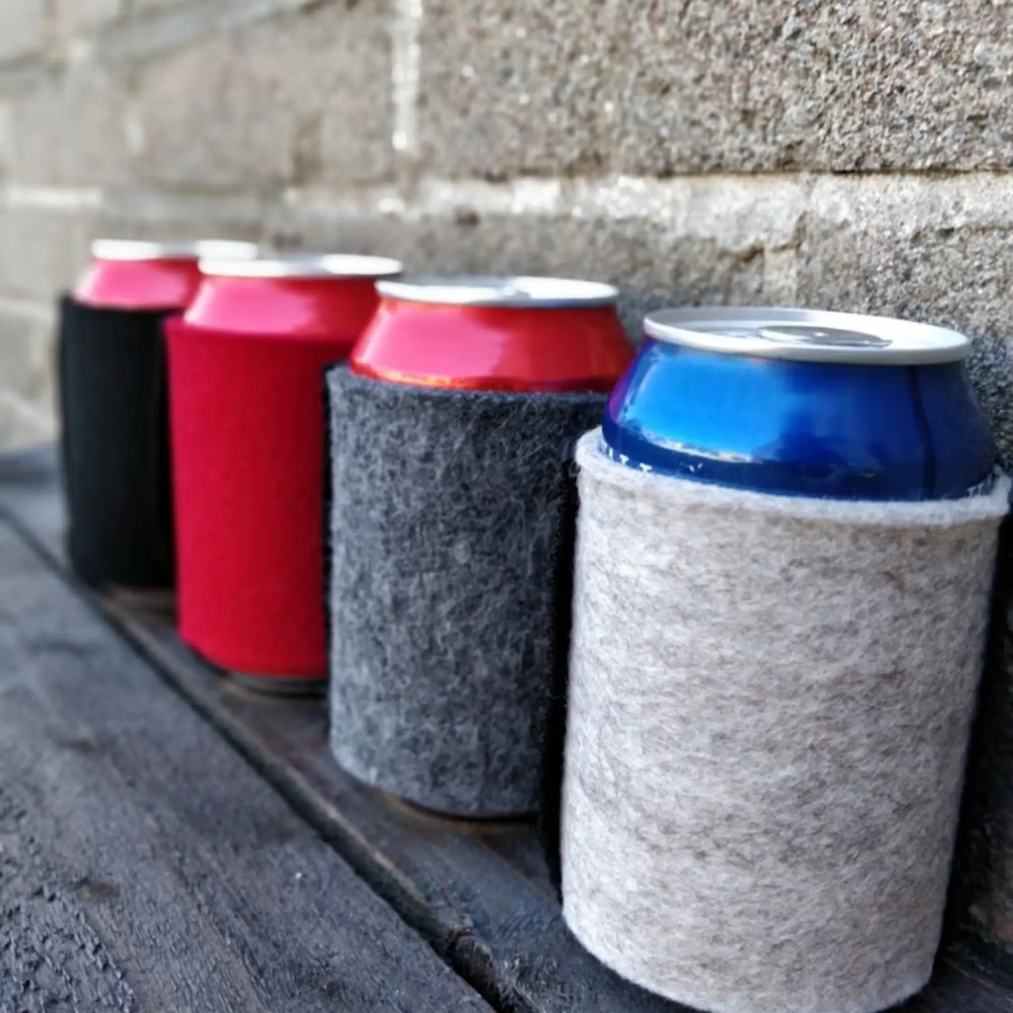 Wool can coolers holding assorted drink cans on a dark wooden bench beside a stone wall.