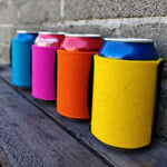 Colourful wool can coolers holding drink cans on a dark wooden bench beside a stone wall.