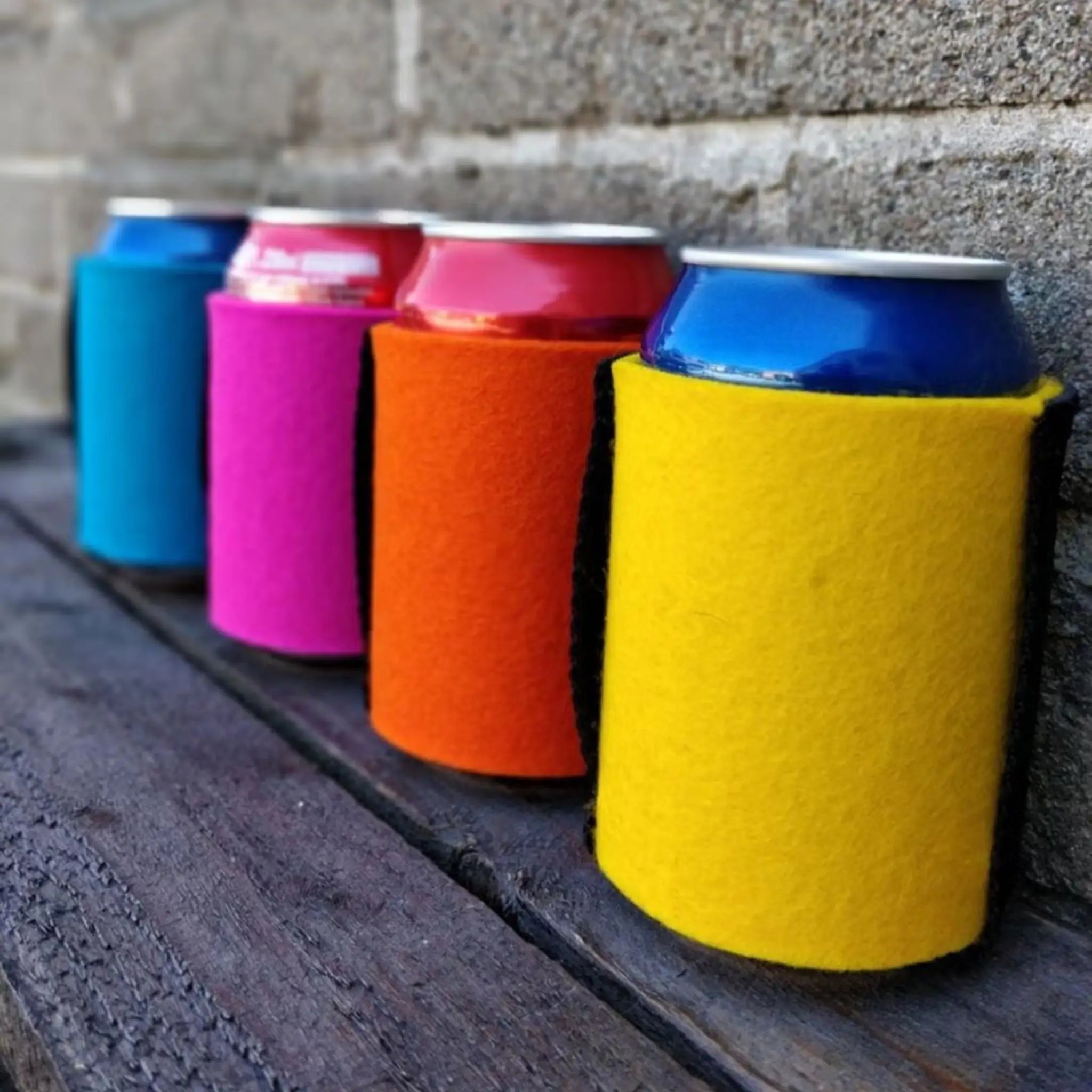 Colourful wool can coolers holding drink cans on a dark wooden bench beside a stone wall.