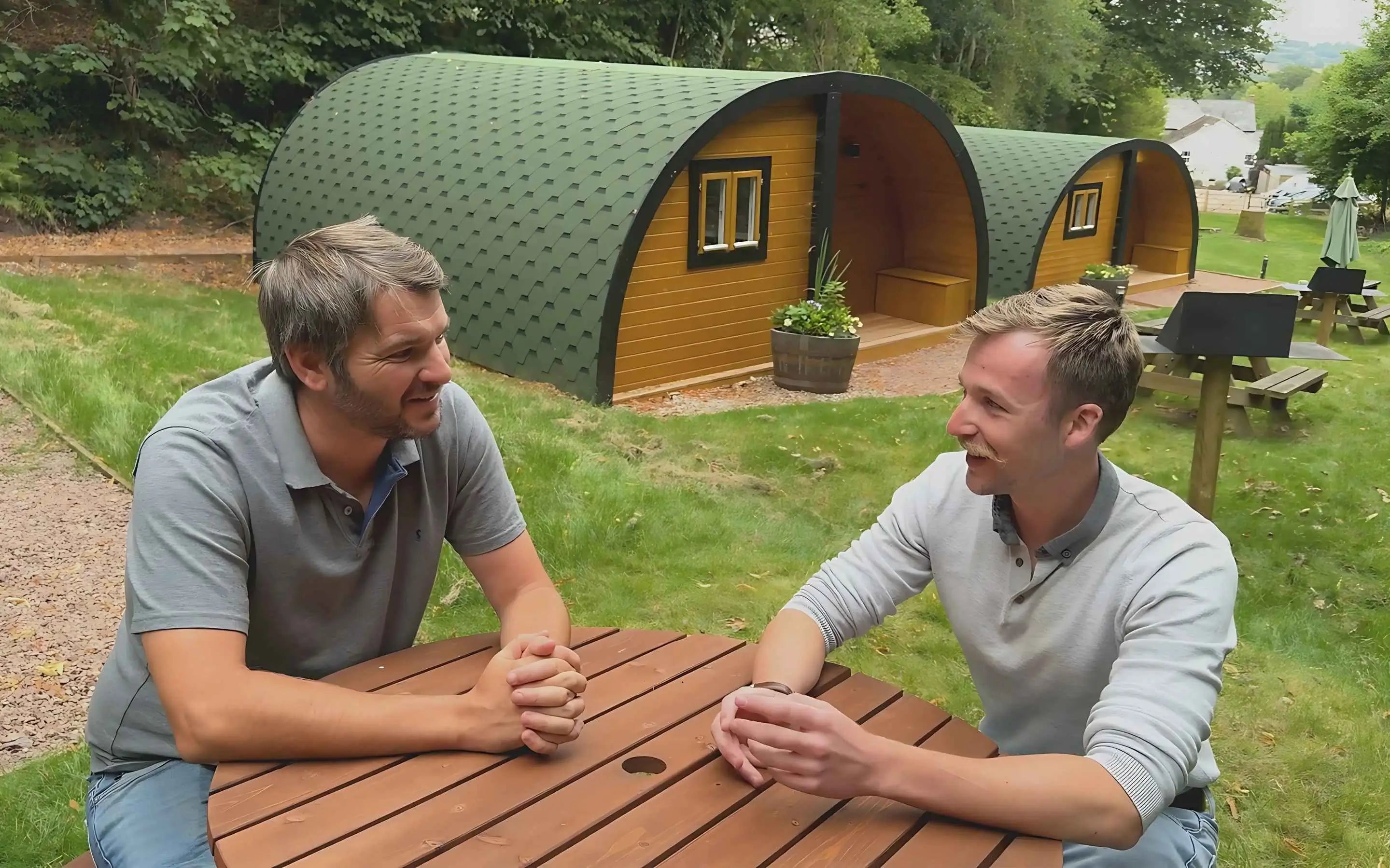 Two men sitting at a wooden table, smiling and talking in front of green-roofed glamping pods at an outdoor site.