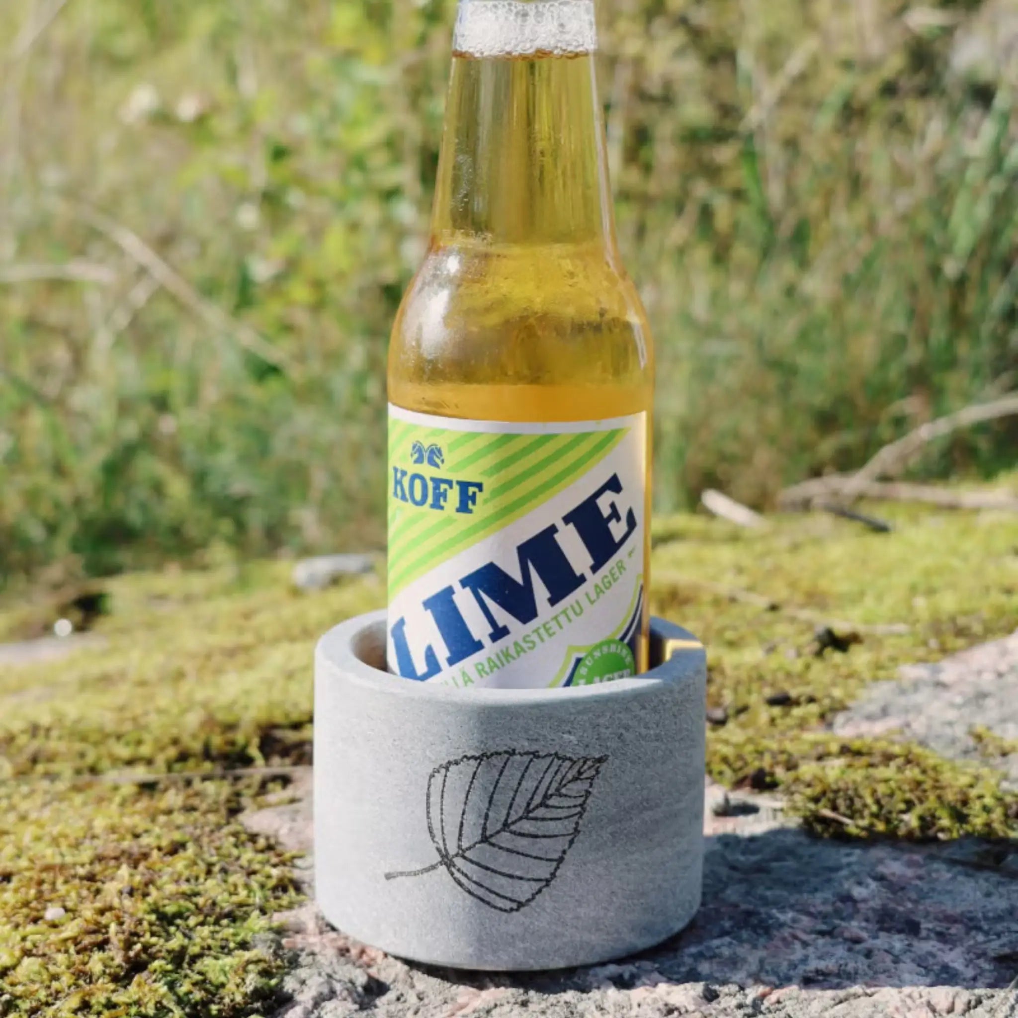 Hukka Saunaviilee soapstone drink cooler holding a glass bottle outdoors on a mossy stone surface.