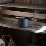Black KOLO sauna pail and ladle placed on a wooden sauna bench inside a traditional sauna room.
