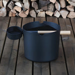 Black KOLO sauna pail and ladle shown beside stacked firewood on a wooden floor.