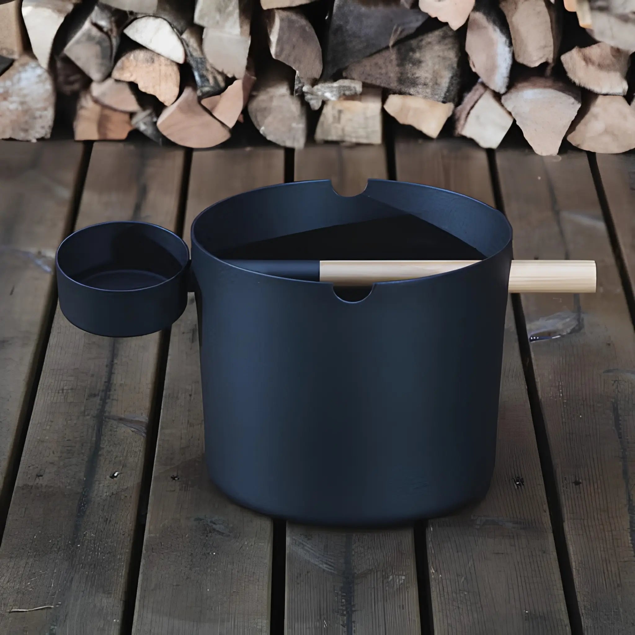 Black KOLO sauna pail and ladle shown beside stacked firewood on a wooden floor.