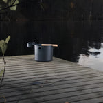 Black KOLO sauna pail and ladle displayed on a wooden lakeside deck beside calm water.