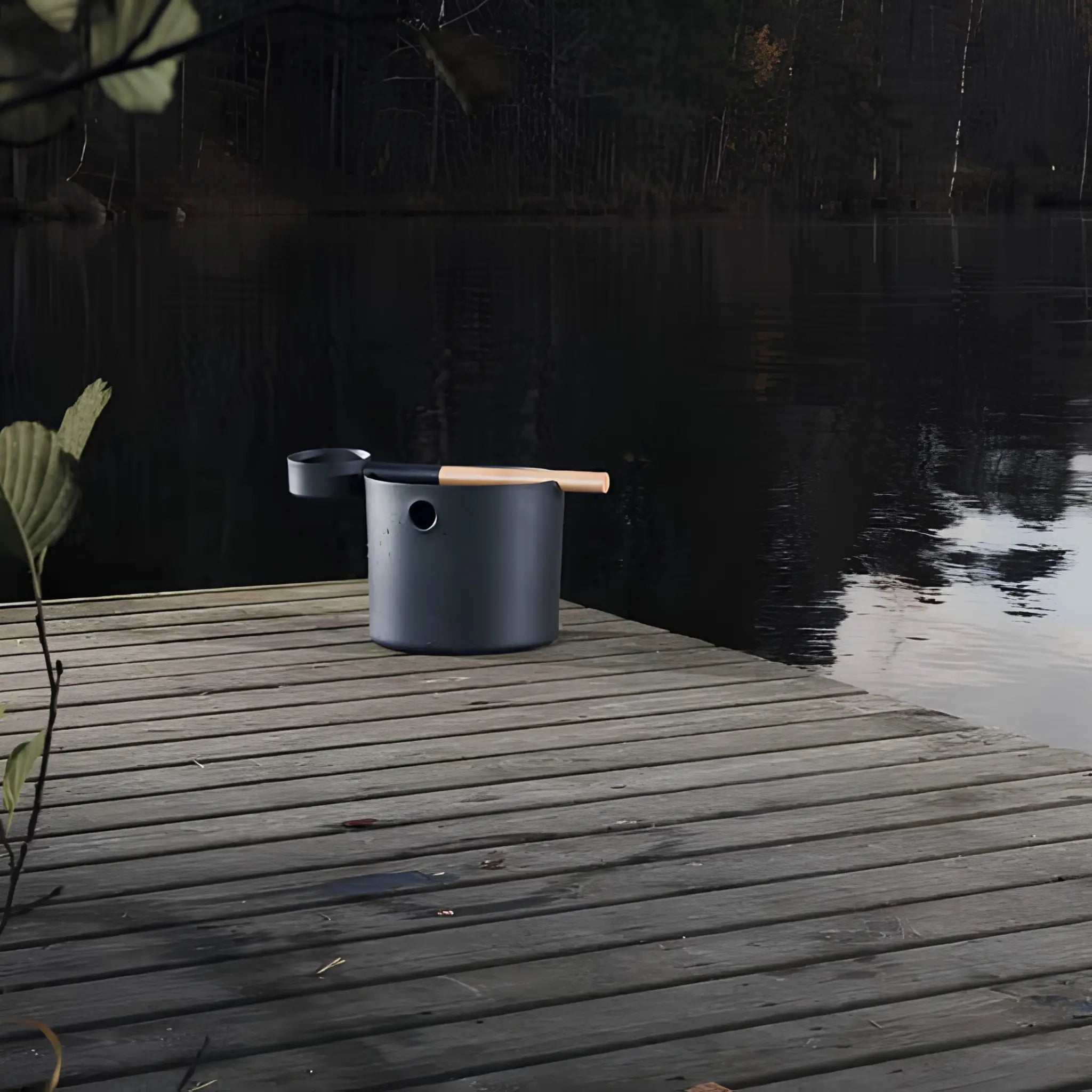 Black KOLO sauna pail and ladle displayed on a wooden lakeside deck beside calm water.