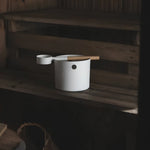 White KOLO sauna pail and ladle placed on a wooden sauna bench inside a softly lit sauna room.