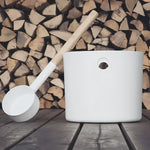 White KOLO sauna pail and ladle styled beside stacked firewood on a wooden sauna floor.