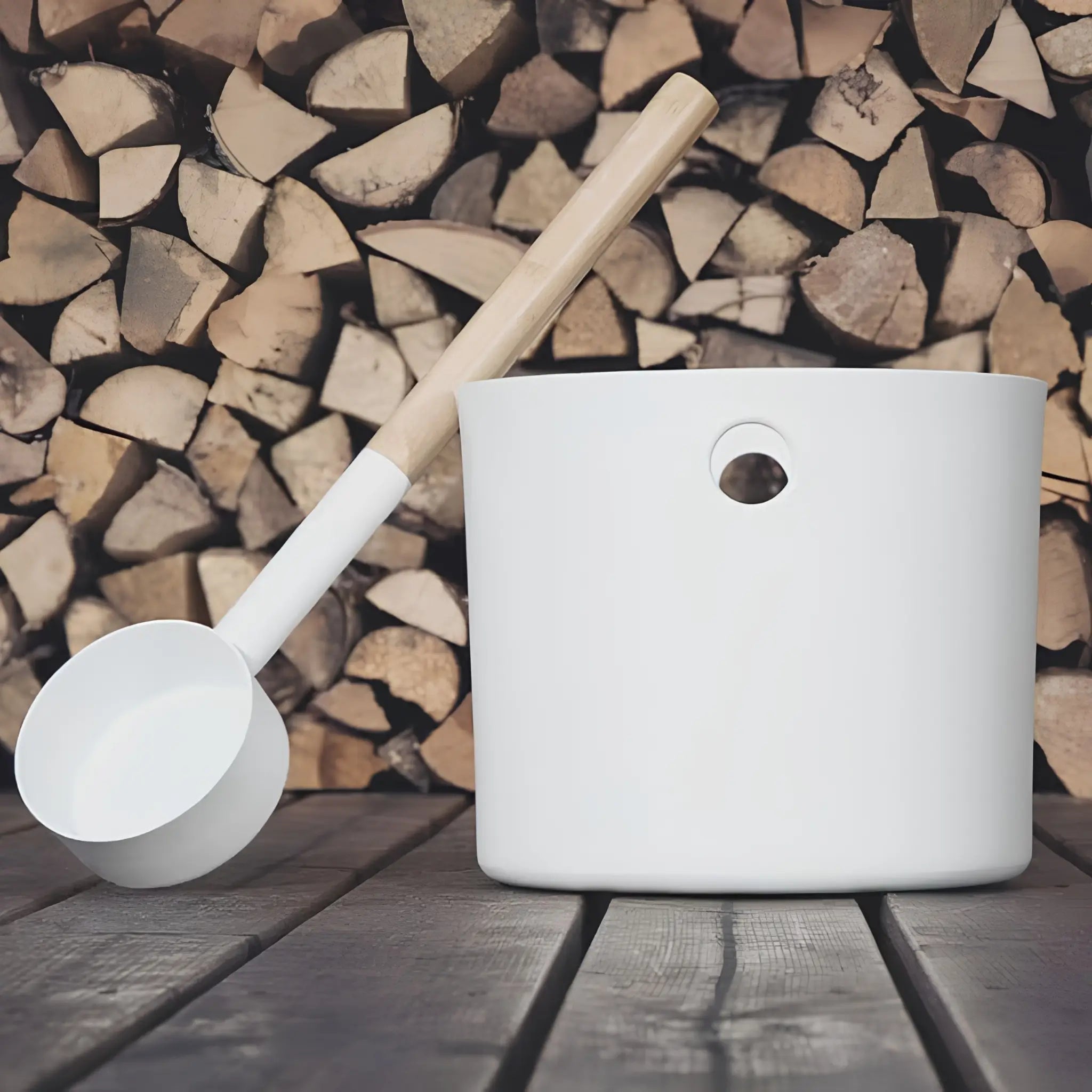 White KOLO sauna pail and ladle styled beside stacked firewood on a wooden sauna floor.