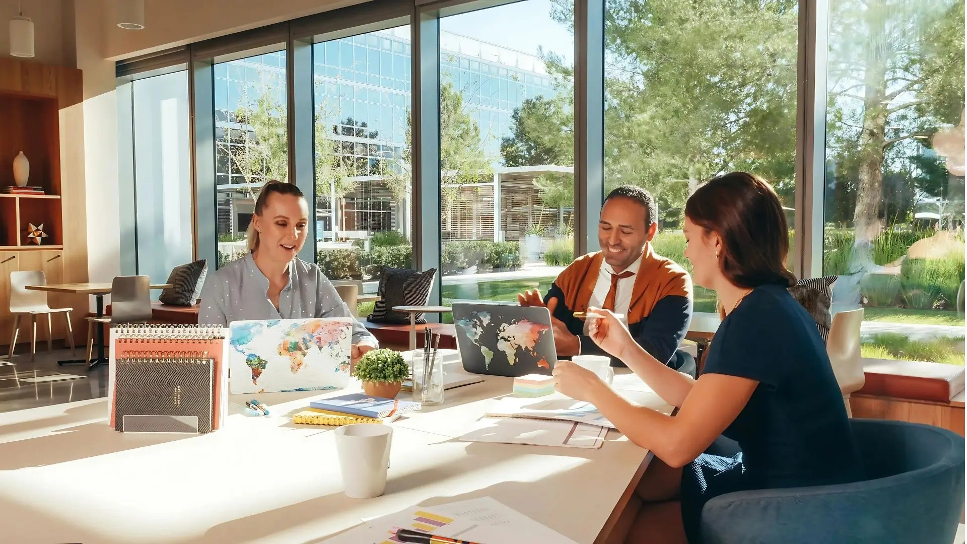 Three professionals collaborating at a table with laptops and notebooks in a sunlit, modern office with large glass windows and greenery outside.