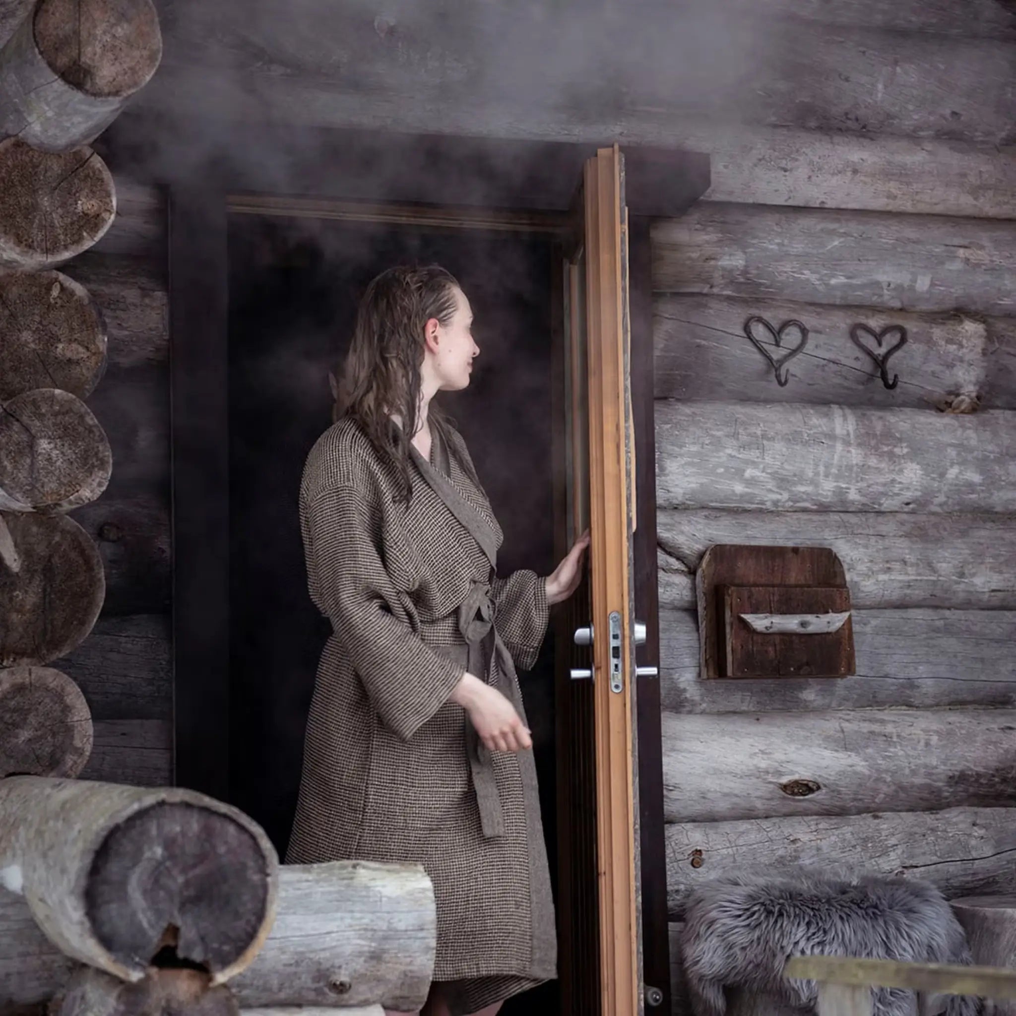Woman wearing a brown Rento Kenno bathrobe outside a rustic wooden sauna cabin.