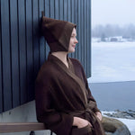 Woman wearing a dark brown Rento Kenno bathrobe and sauna hat outside a winter sauna cabin.