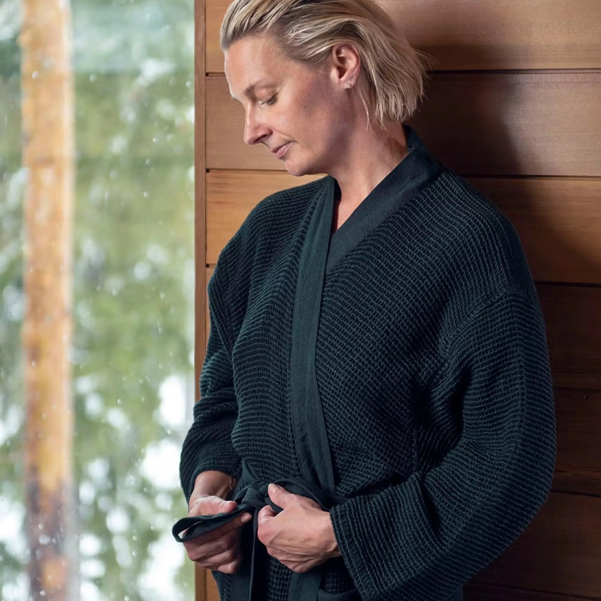 Woman tying the belt of a dark green Rento Kenno bathrobe in a sauna cabin setting.