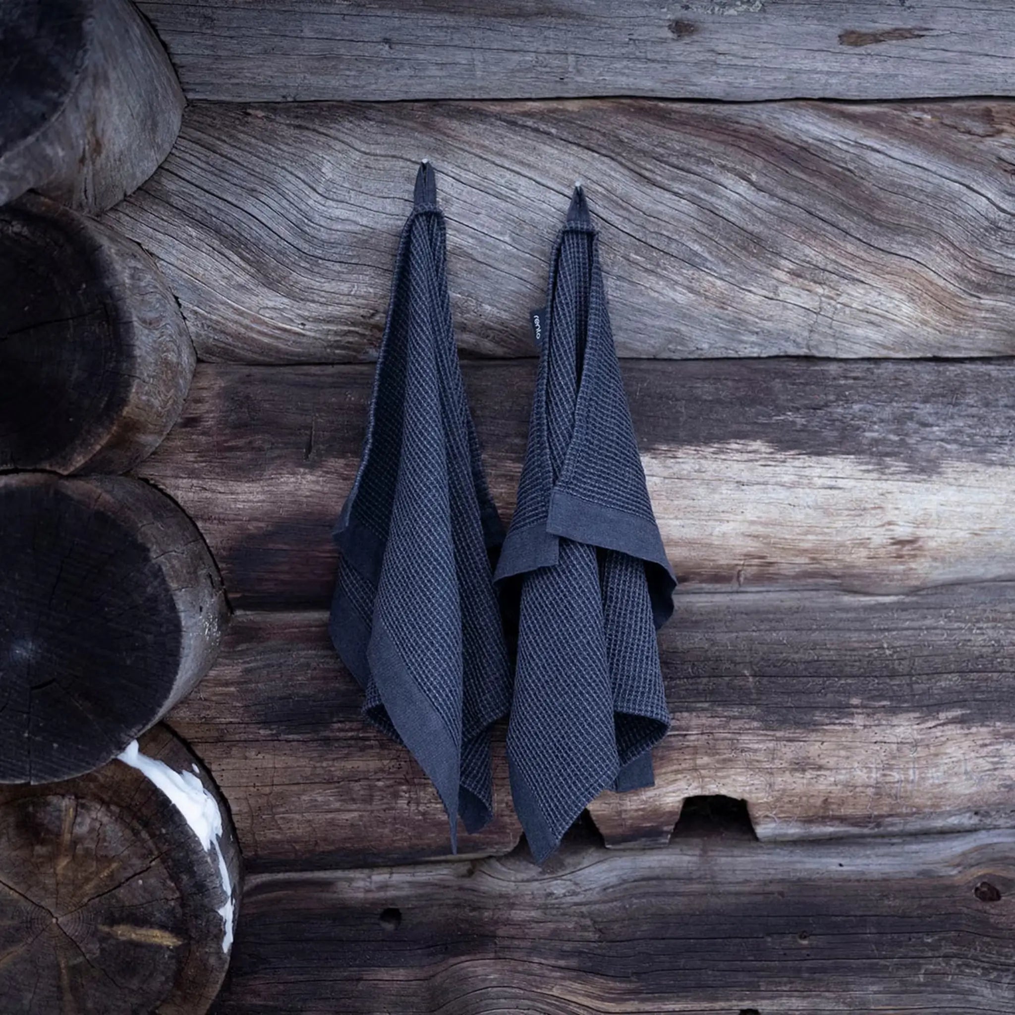 Black Rento Kenno hand towels hanging on a log wall in a traditional sauna setting.