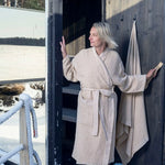 Beige waffle bath towel hanging beside a sauna doorway with a model wearing a matching beige bathrobe in a snowy outdoor sauna setting