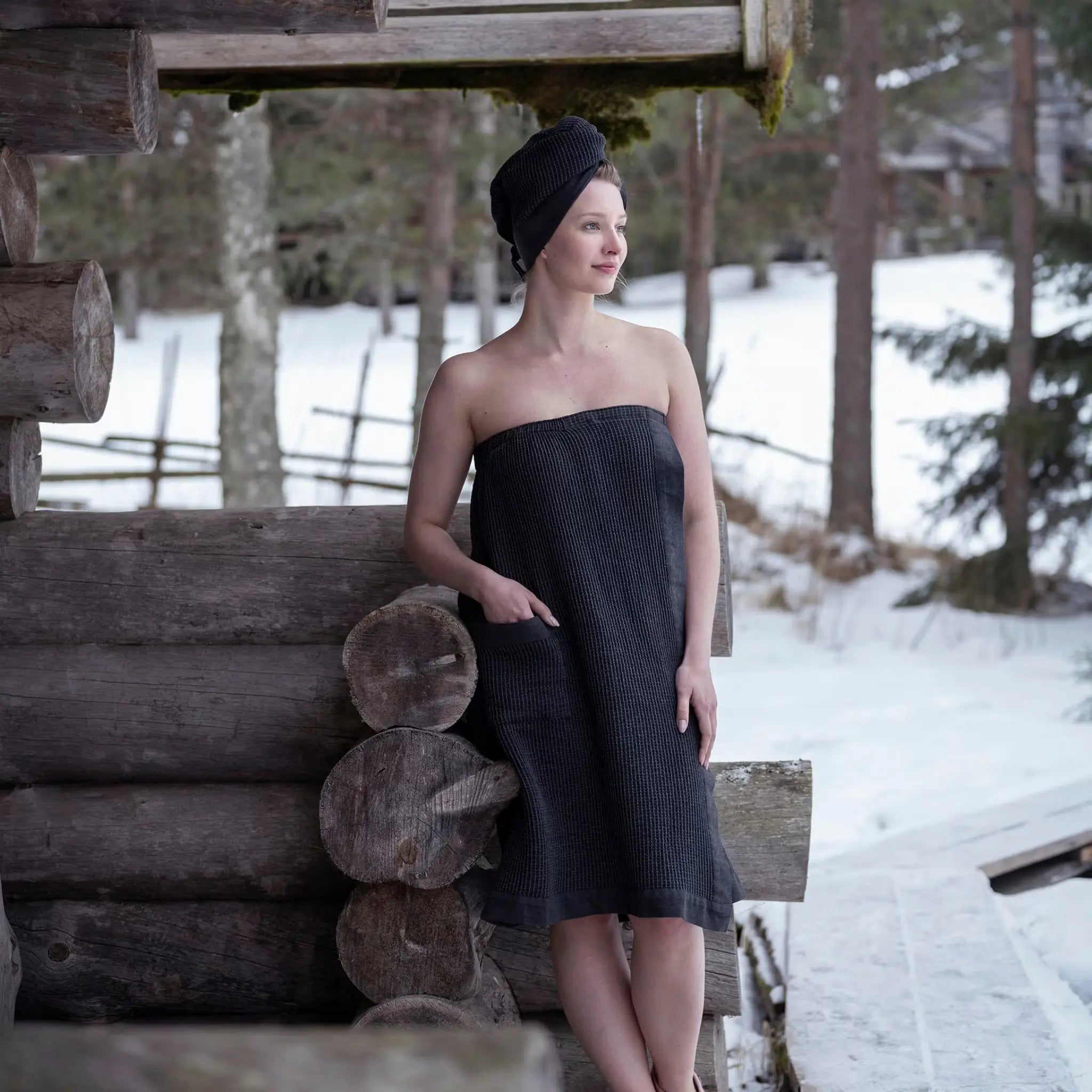 Woman wearing a black sauna sarong wrap towel outside a rustic sauna cabin in snowy surroundings