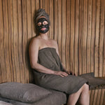Woman seated in brown sauna wrap towel with face mask and natural sauna accessories
