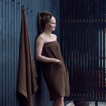 Woman relaxing in a dark brown sauna sarong beside a hanging towel on a contemporary outdoor sauna deck