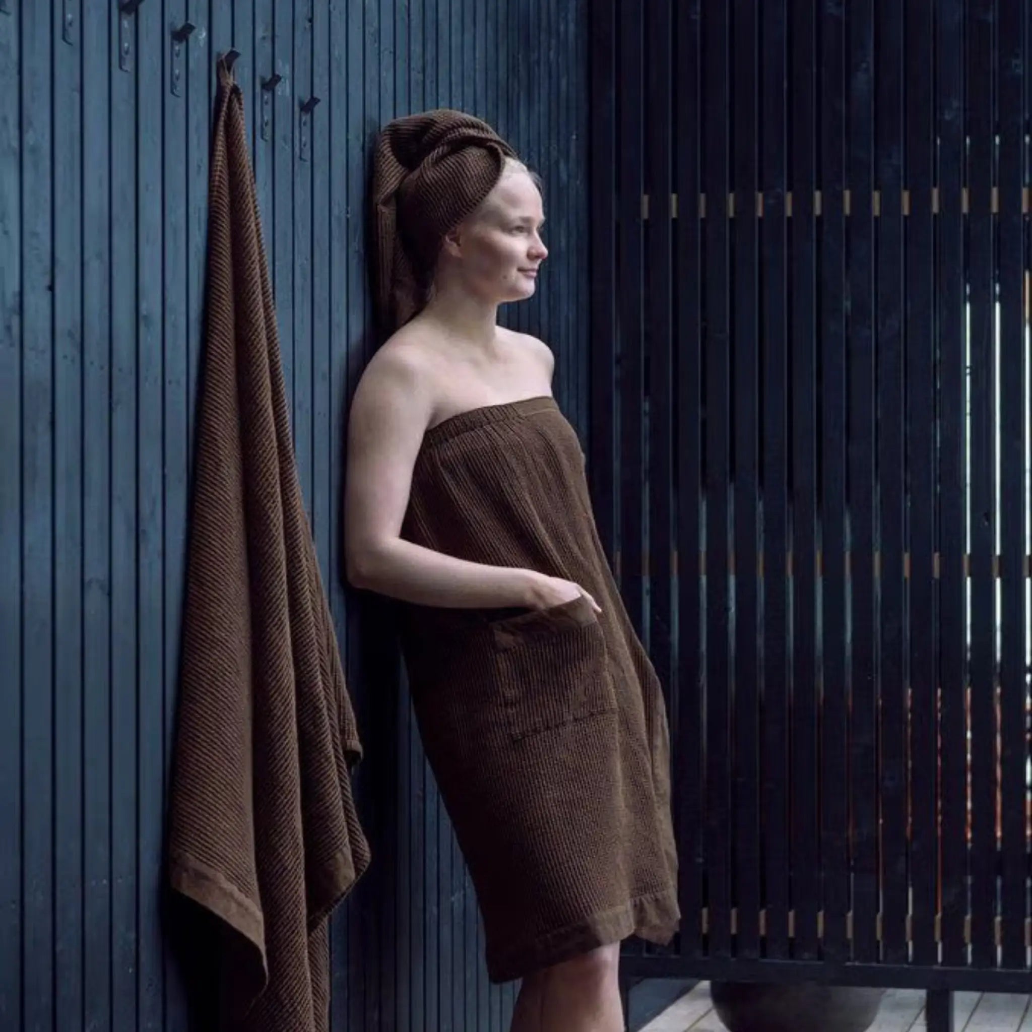 Woman relaxing in a dark brown sauna sarong beside a hanging towel on a contemporary outdoor sauna deck