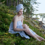 Woman relaxing in a grey sauna sarong and matching hair towel on mossy ground beside a calm lake.