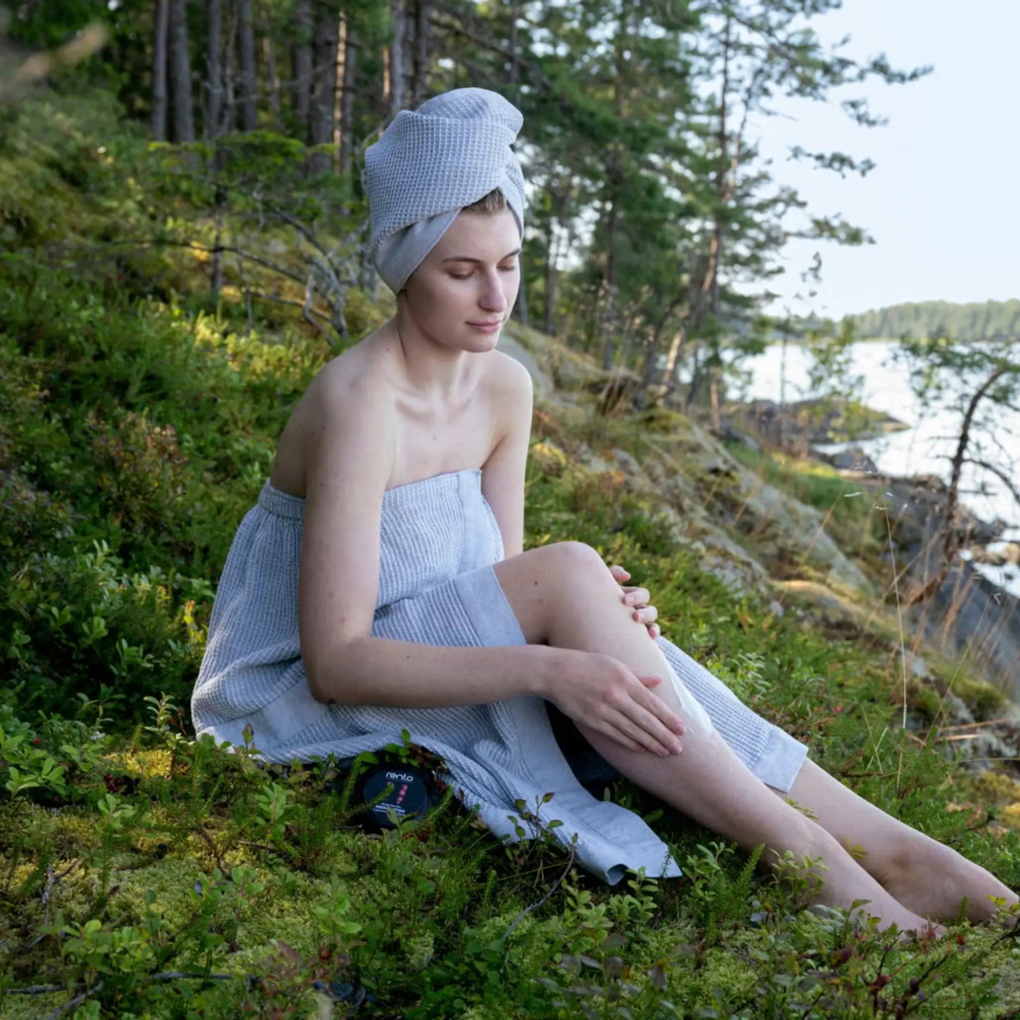 Woman relaxing in a grey sauna sarong and matching hair towel on mossy ground beside a calm lake.