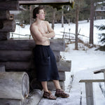 Man wearing a black waist towel outside a log sauna in a snowy forest setting