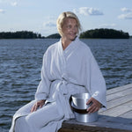 Woman wearing a grey Rento Kenno bathrobe while sitting on a lakeside deck with a sauna bucket.