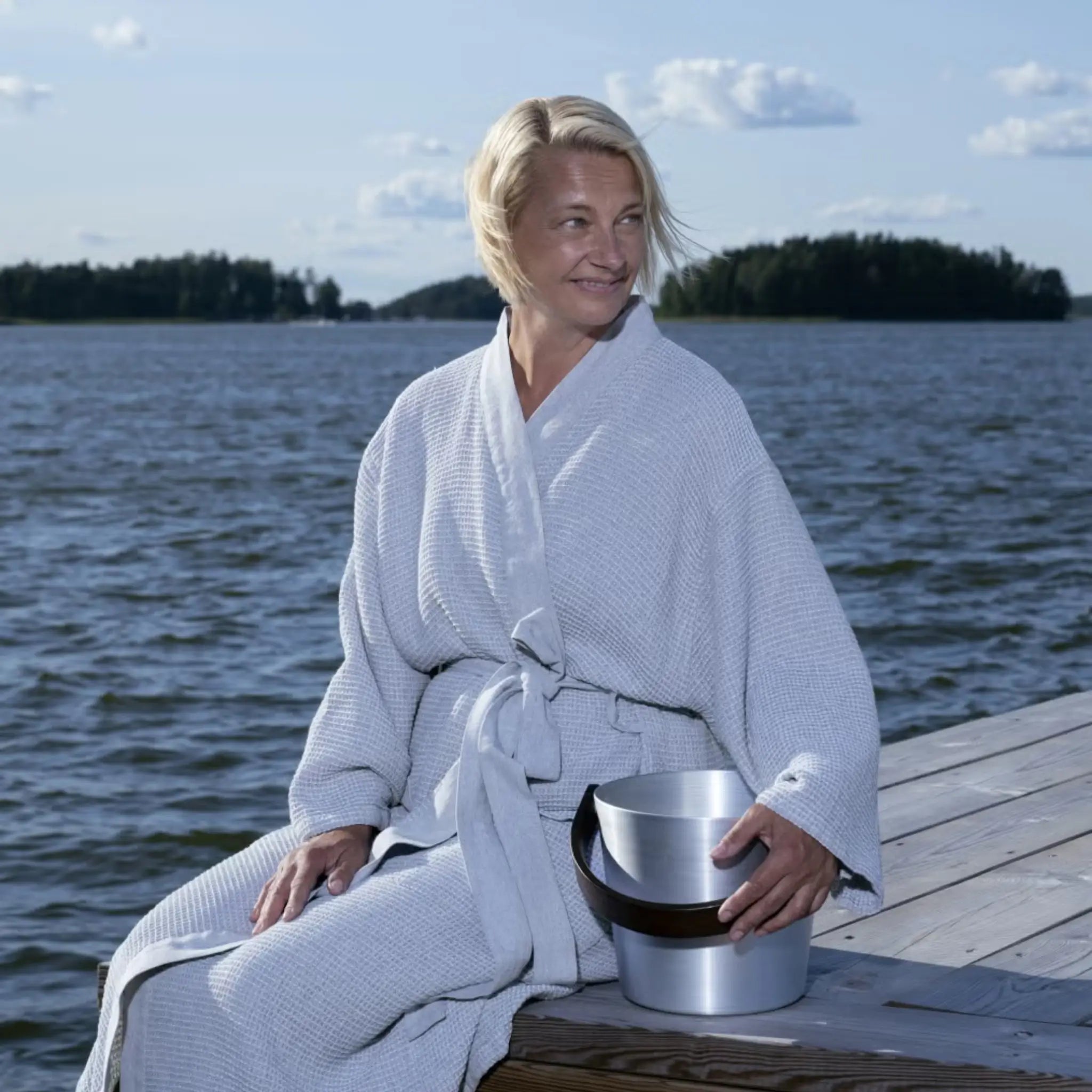 Woman wearing a grey Rento Kenno bathrobe while sitting on a lakeside deck with a sauna bucket.