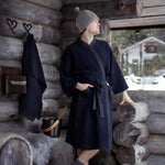 Man wearing a black Rento Kenno bathrobe inside a rustic log cabin with sauna accessories nearby.