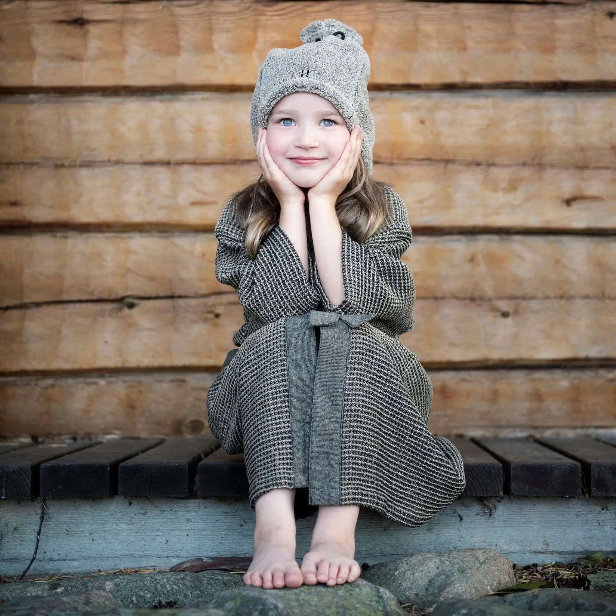 Child sitting outdoors in a brown Rento Kenno kids bathrobe with matching belt and textured fabric