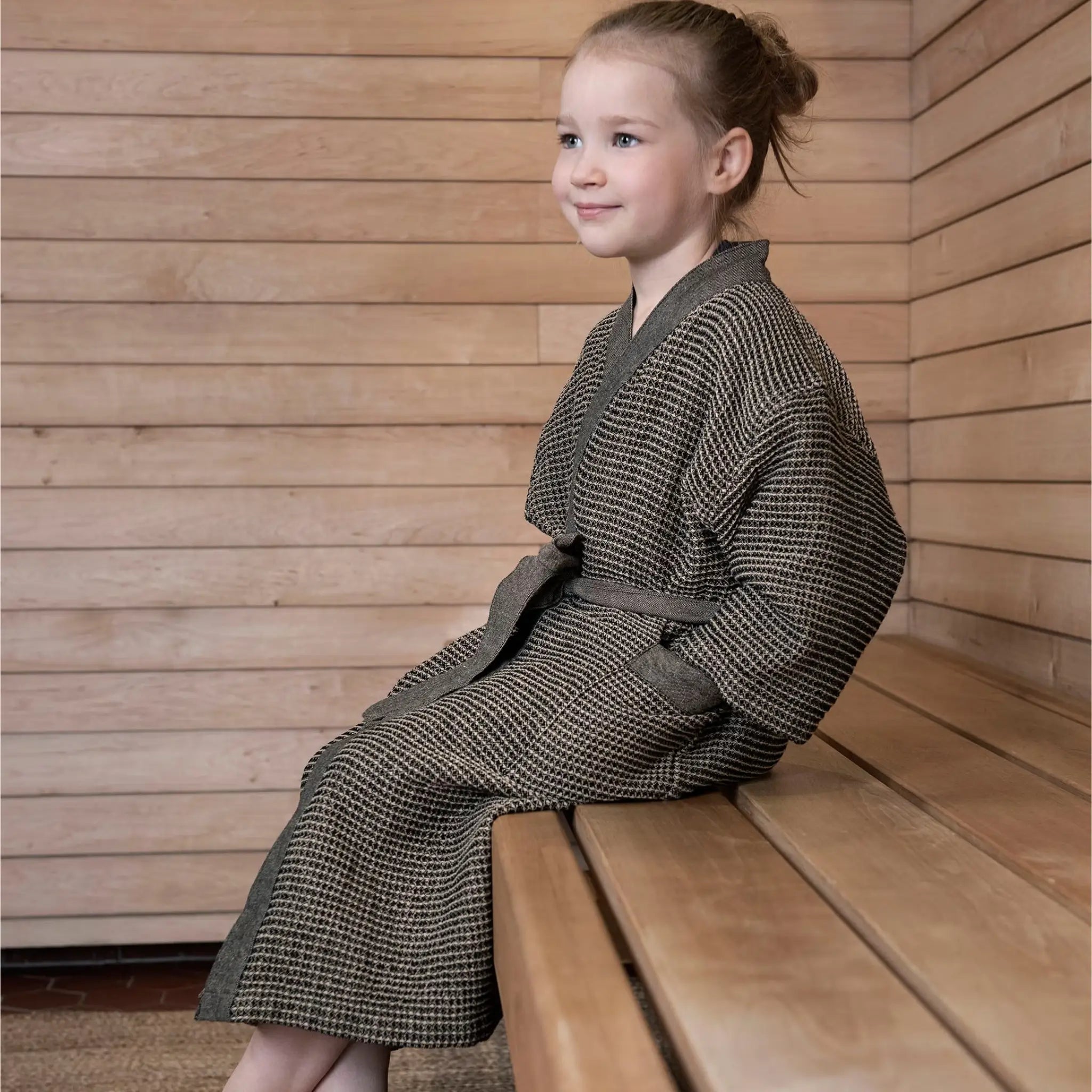 Child wearing a brown Rento Kenno kids bathrobe while sitting on a wooden sauna bench