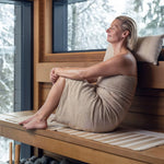 Woman wearing a beige sauna towel outdoors by a lake while holding a sauna bucket and ladle