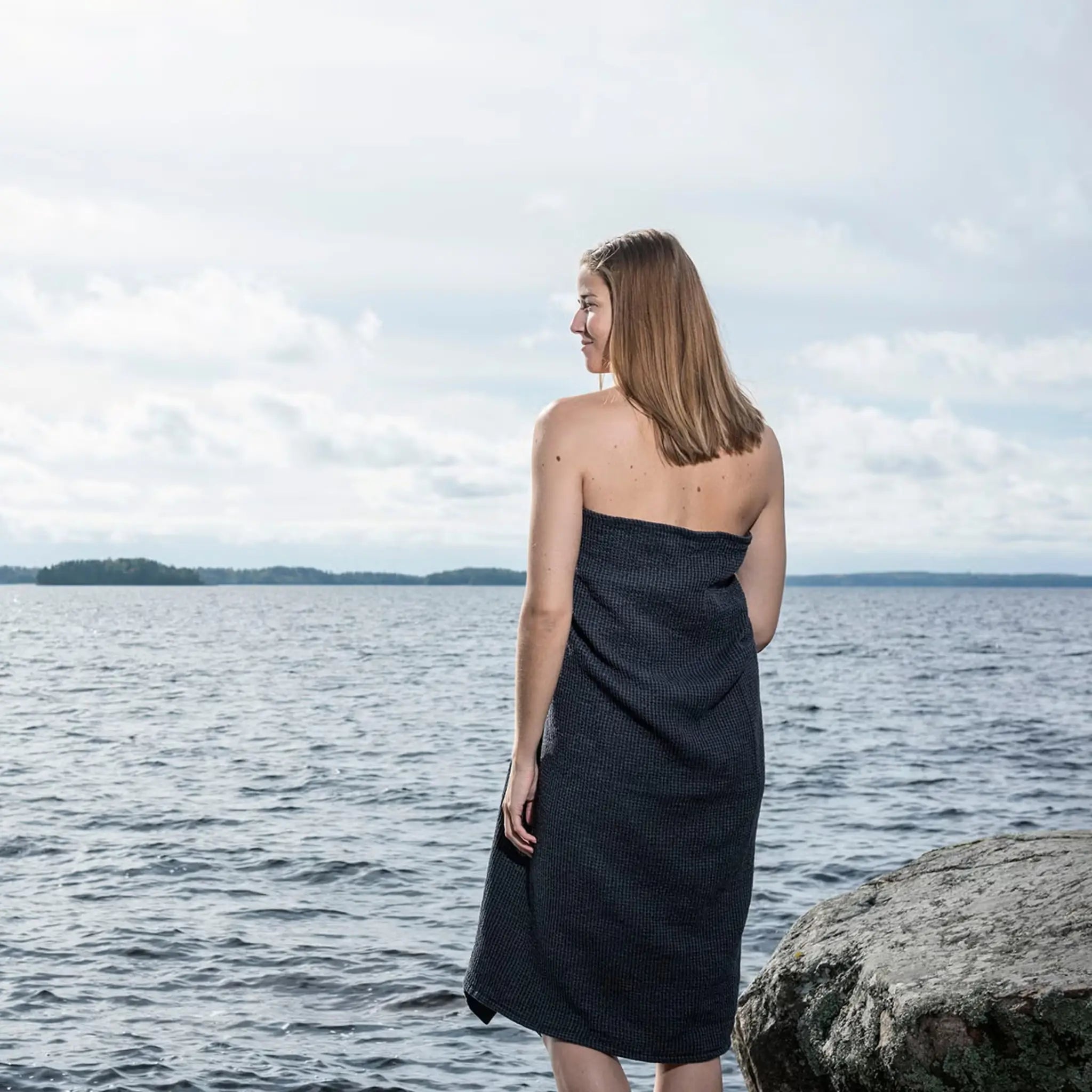Black large bath towel worn as a sauna wrap by a woman standing beside a lake