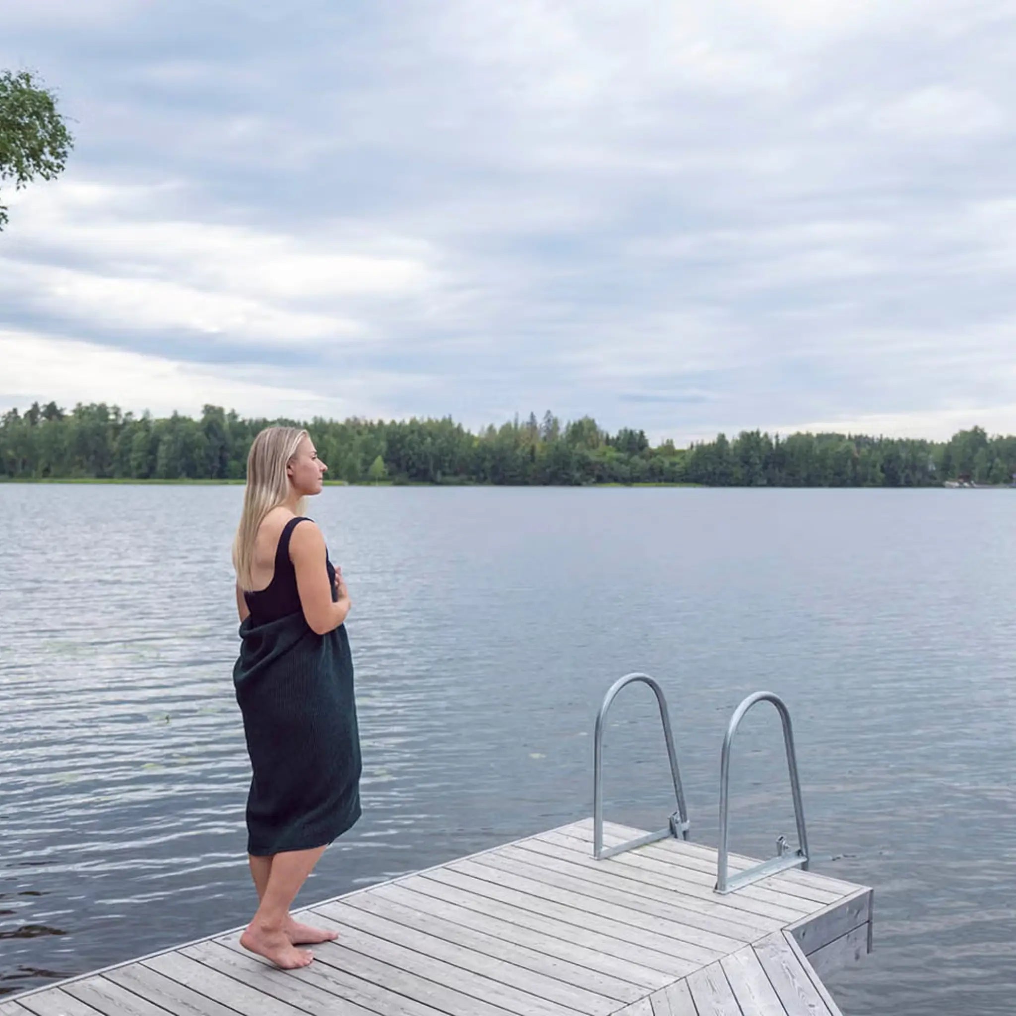 Woman wrapped in a dark green waffle texture bath towel standing on a wooden jetty by a lake
