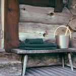 Dark green bath towels stacked on a rustic wooden bench beside a sauna bucket