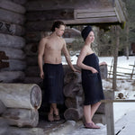 Couple wearing black sauna wraps and slippers outside a rustic log sauna in winter