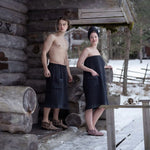 Couple in black sauna wraps standing outside a snow-covered log sauna cabin