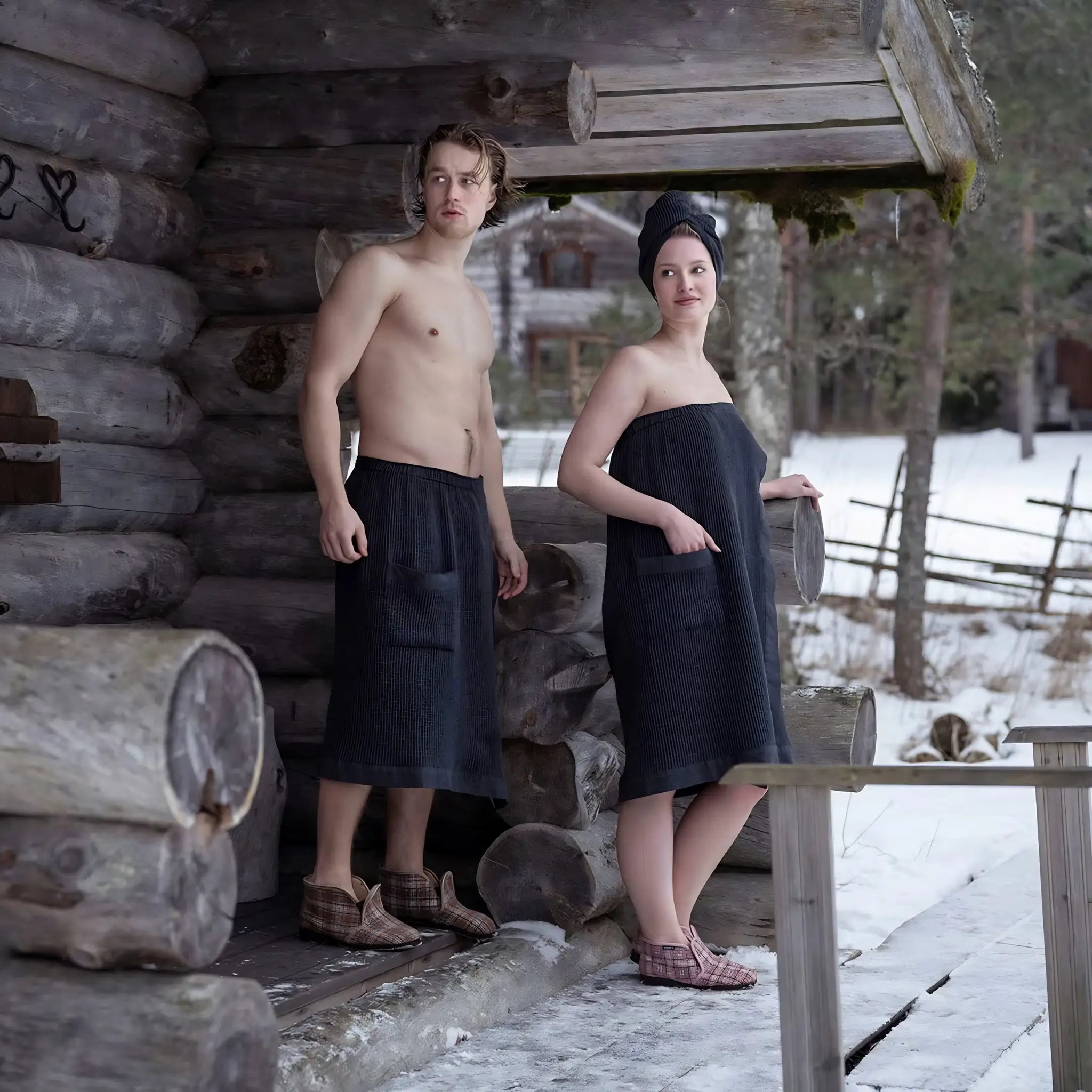 Couple in black sauna wraps standing outside a snow-covered log sauna cabin