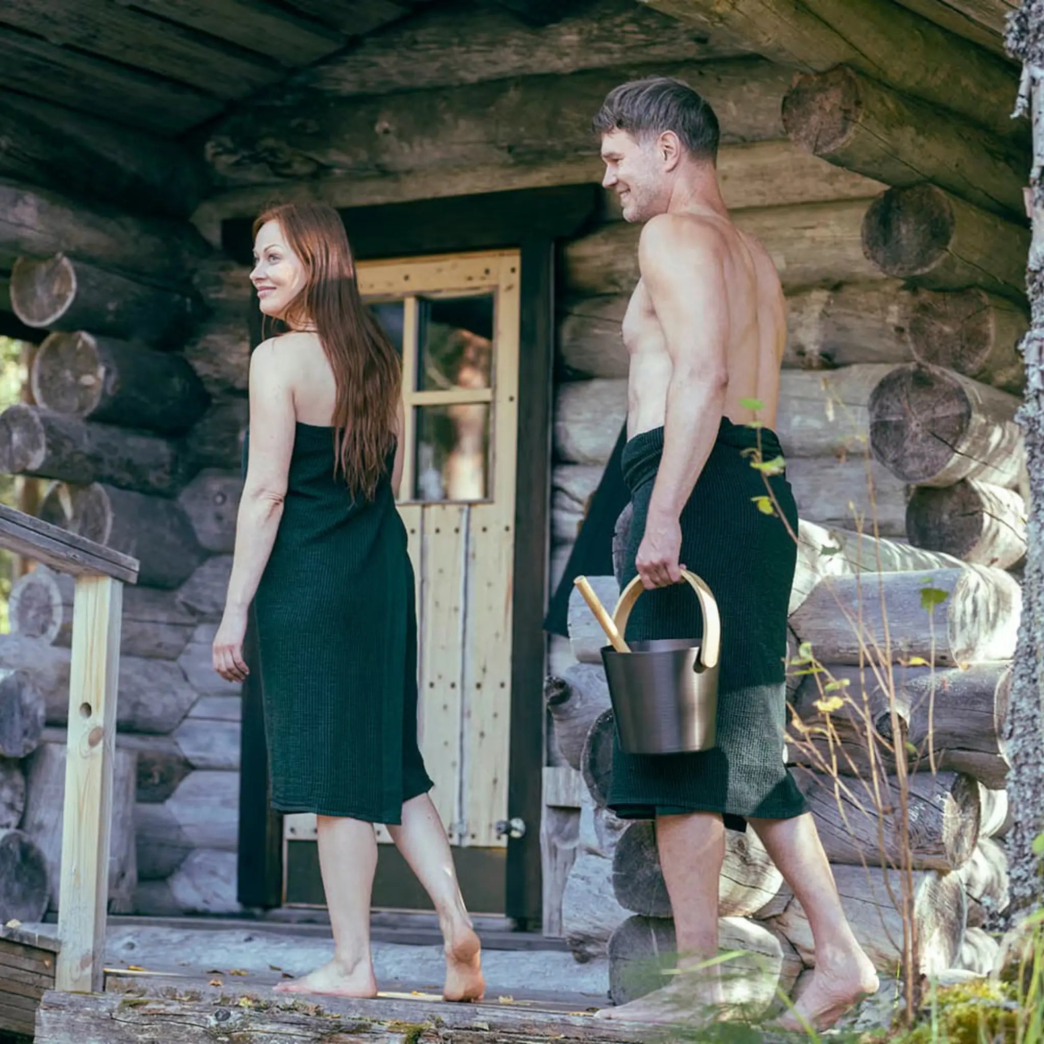 Couple walking outside a log sauna wearing dark green matching sauna wraps