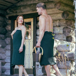 Couple wearing dark green sauna wraps outside a rustic sauna cabin with bucket and ladle