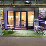 Fully furnished Small GlampTainer container-style glamping structure – illuminated front view featuring cosy bedroom, kitchenette, and outdoor seating setup.