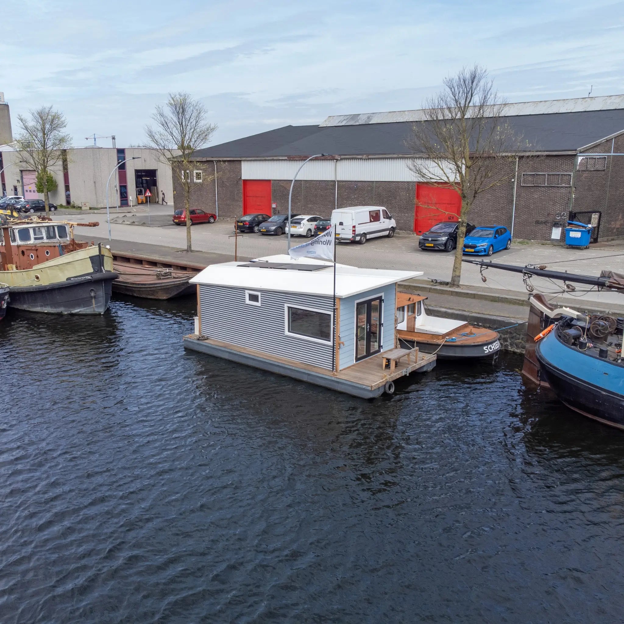 Side aerial view of a floating shepherd’s hut moored between boats on a calm waterfront canal.